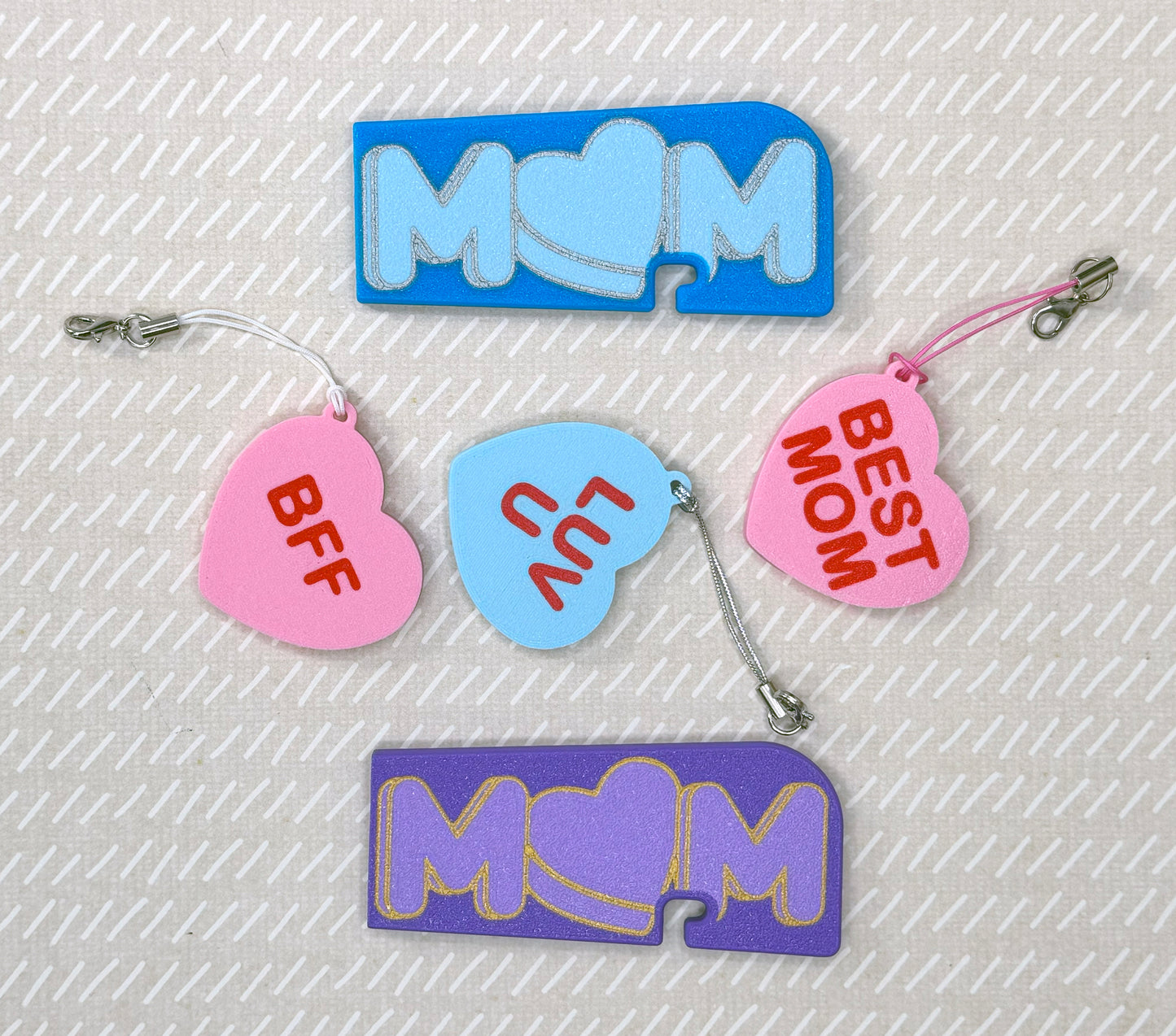 Various colorful heart and Mom-themed soda Tab Buddies and Tab Buddy XLs on a textured beige background