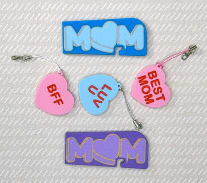 Various colorful heart and Mom-themed soda Tab Buddies and Tab Buddy XLs on a textured beige background