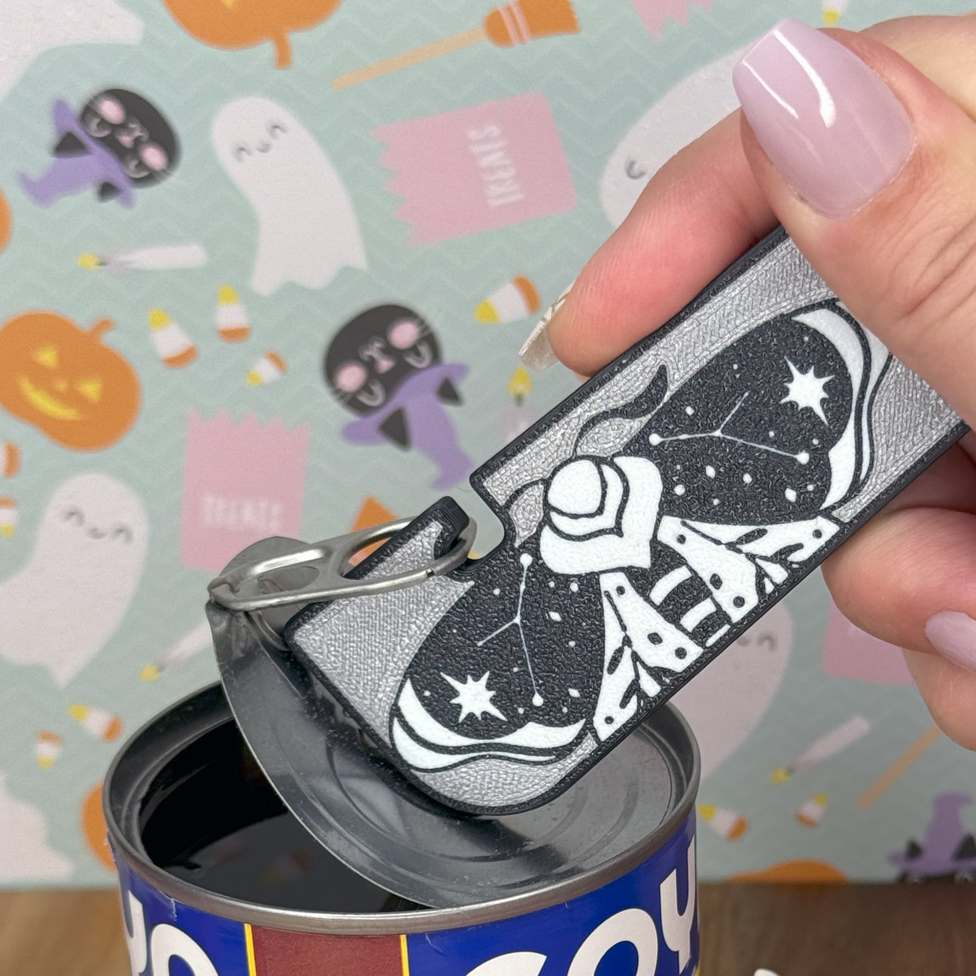 Hand using a silver, black and white moth-designed Tab Buddy XL to open a can with a colorful background featuring ghosts and pumpkins.