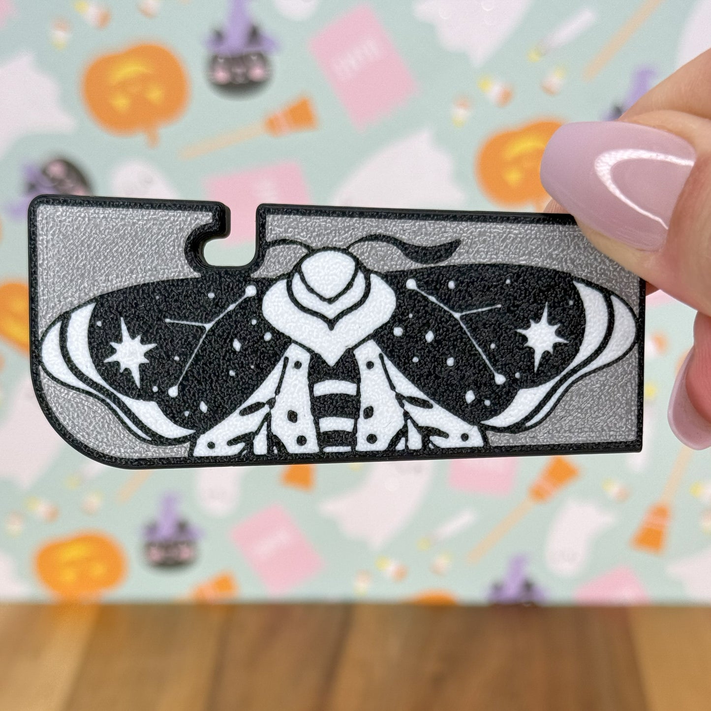 Decorative moth design Tab Buddy XL held by a hand with pink nail polish against a colorful blurred background.