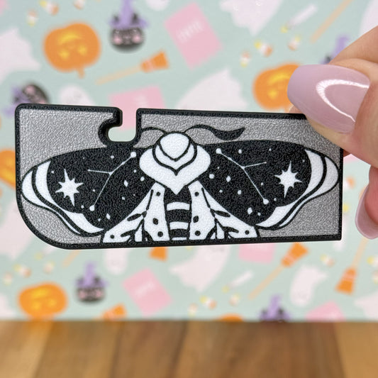 Decorative moth design Tab Buddy XL held by a hand with pink nail polish against a colorful blurred background.