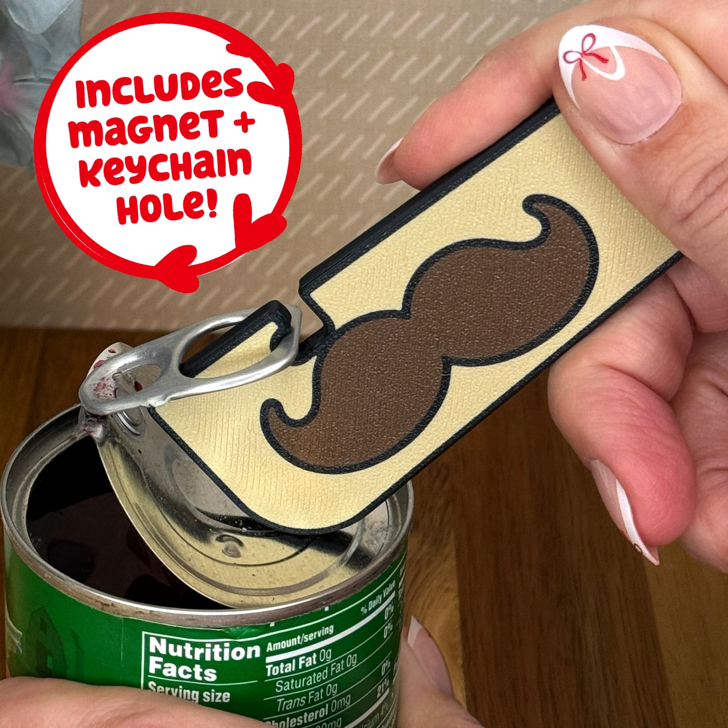 Mustache-themed Tab Buddy XL is used to open a can of vegetables. Text notes the Tab Buddy XL includes a magnet and keychain hole.