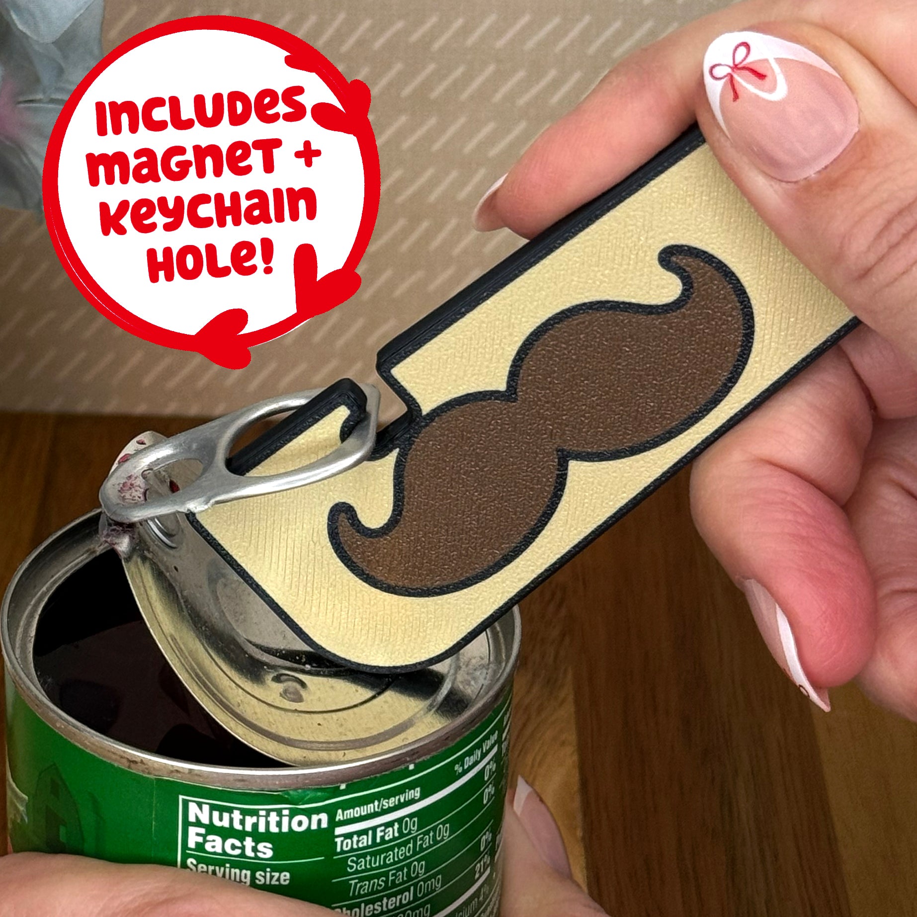 Mustache-themed Tab Buddy XL is used to open a can of vegetables. Text notes the Tab Buddy XL includes a magnet and keychain hole.