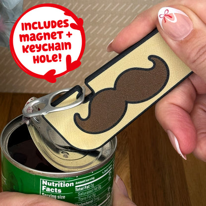 Mustache-themed Tab Buddy XL is used to open a can of vegetables. Text notes the Tab Buddy XL includes a magnet and keychain hole.