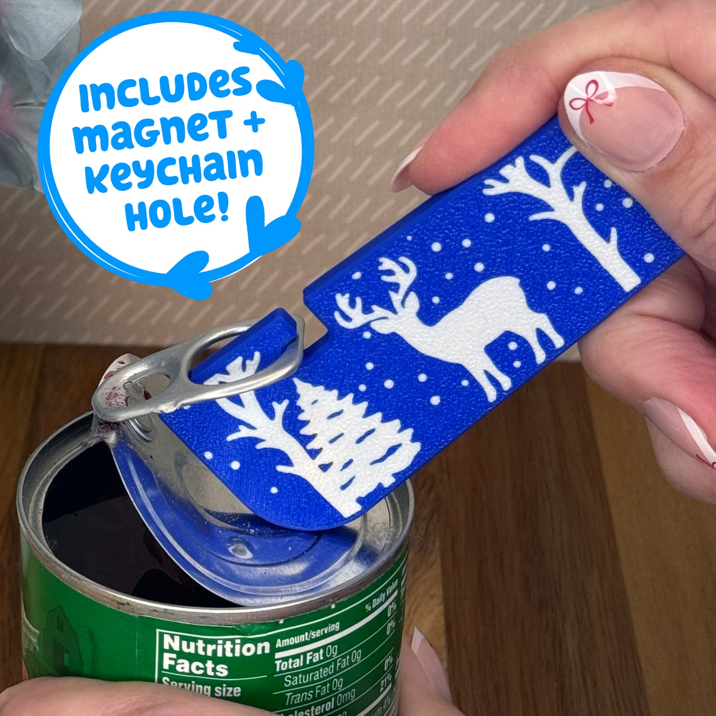 Person using a Tab Buddy XL with a blue and white reindeer design on a can. Text notes the Tab Buddy XL includes a magnet and keychain hole.