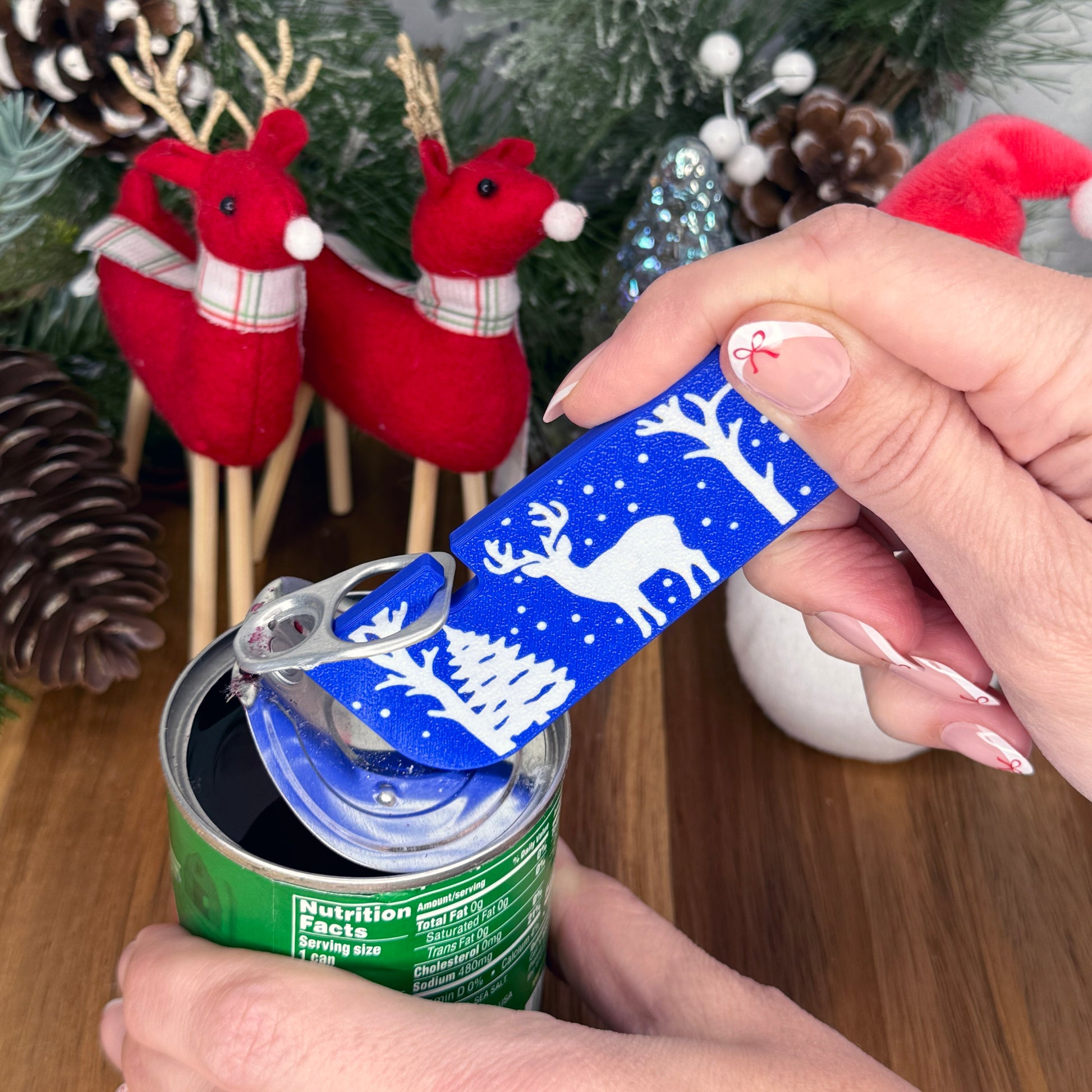 Hand using a blue reindeer-themed Tab Buddy XL on a green can with Christmas decorations in the background.
