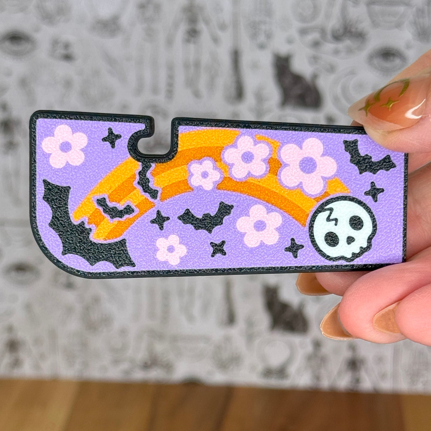 Decorative Tab Buddy XL with Halloween-themed orange rainbow, bat and skull design held by a hand against a neutral background