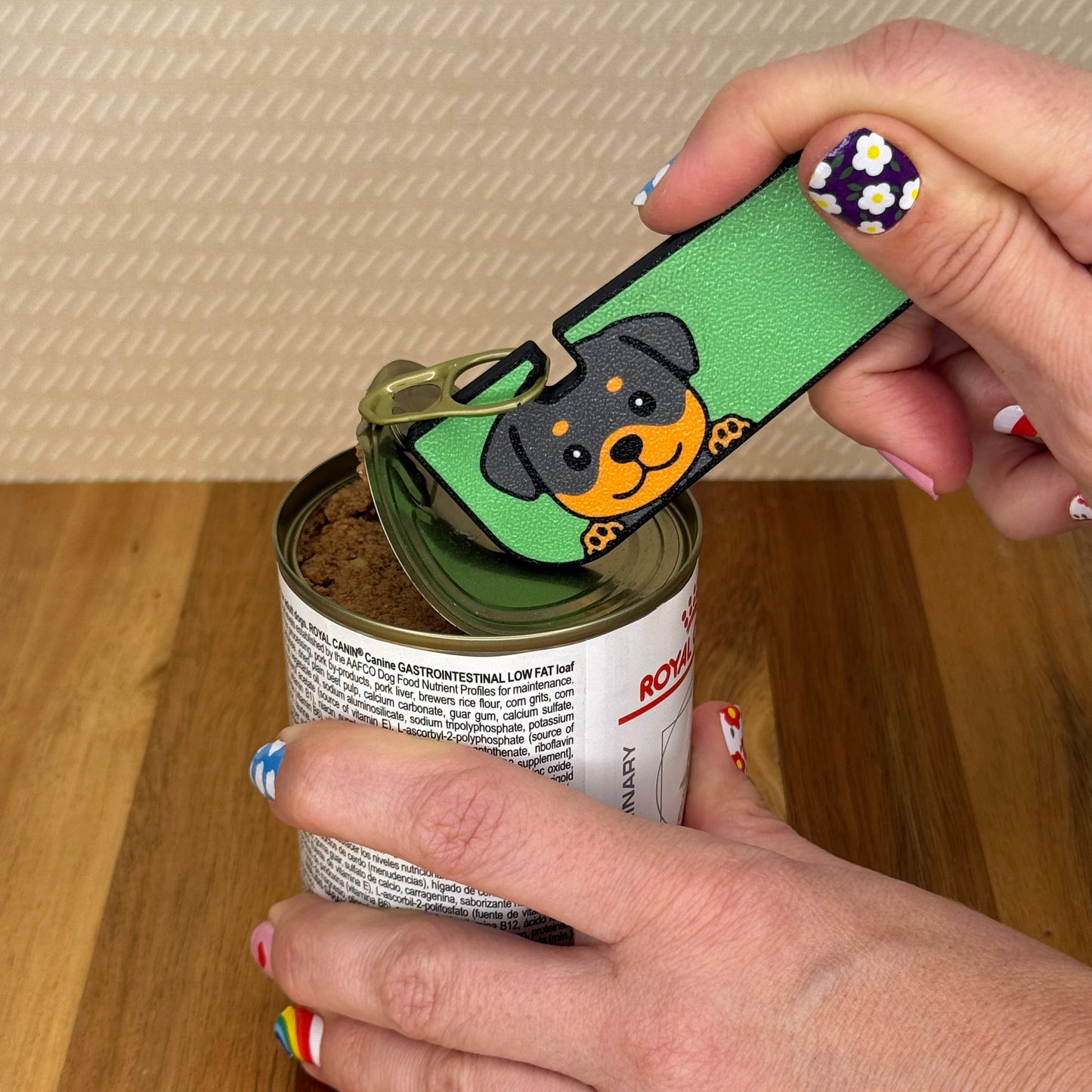 Hand using a Tab Buddy XL with a Rottweiler dog design to open a can of dog food on a wooden surface.