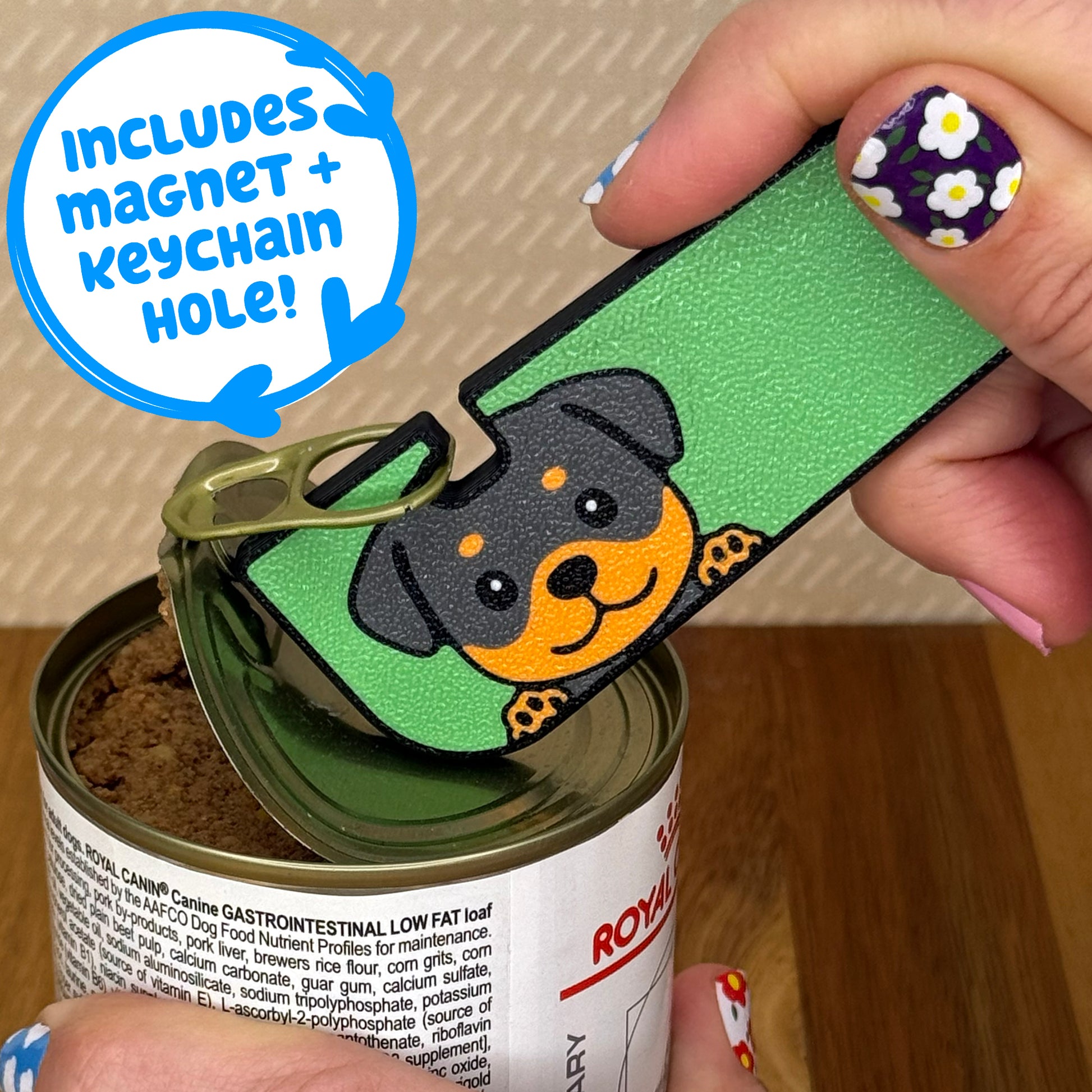 Hand using a Tab Buddy XL with a Rottweiler dog design to open a can of dog food on a wooden surface. Text bubble reads, "Includes magnet + keychain strap!"