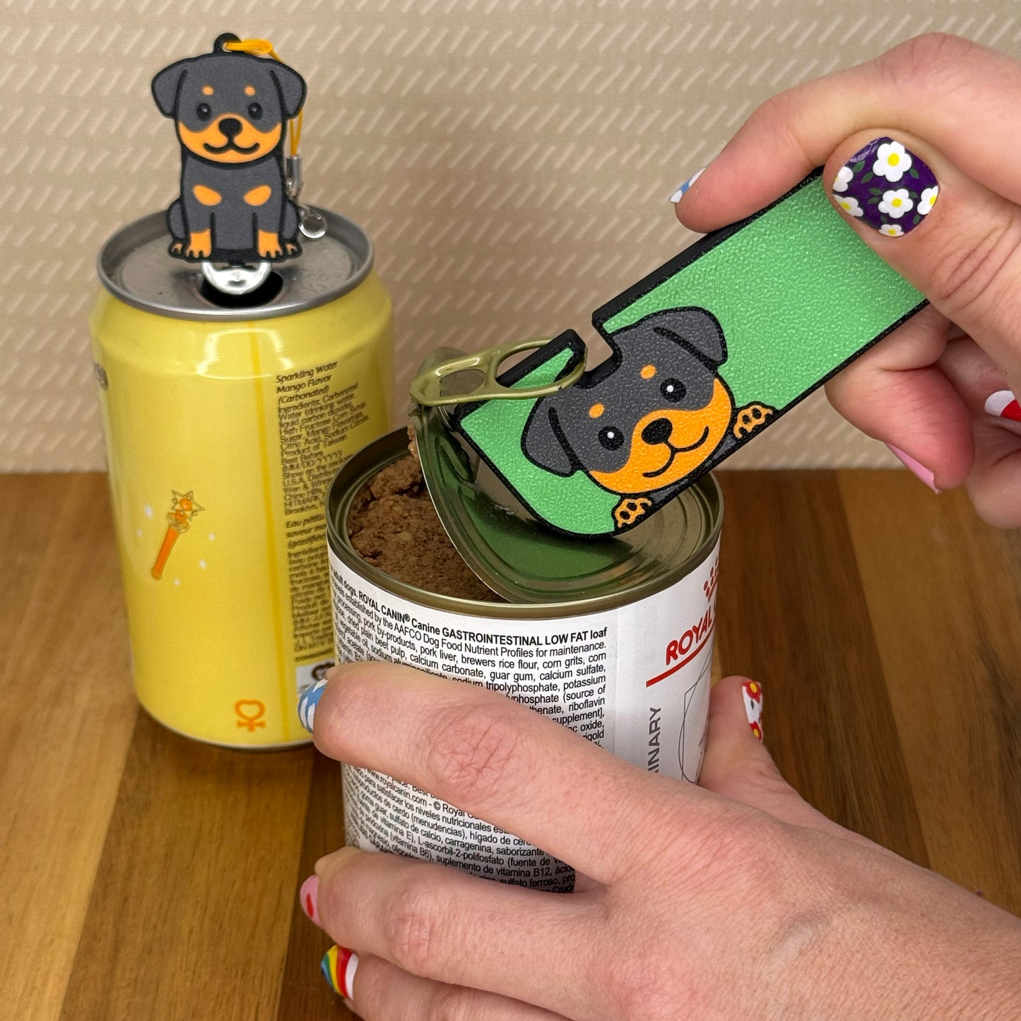 A person opens a can of dog food with a Rottweiler Dog-design Tab Buddy XL. In the background, a matching soda-sized Tab Buddy rests atop the tab of a can of sparkling water.