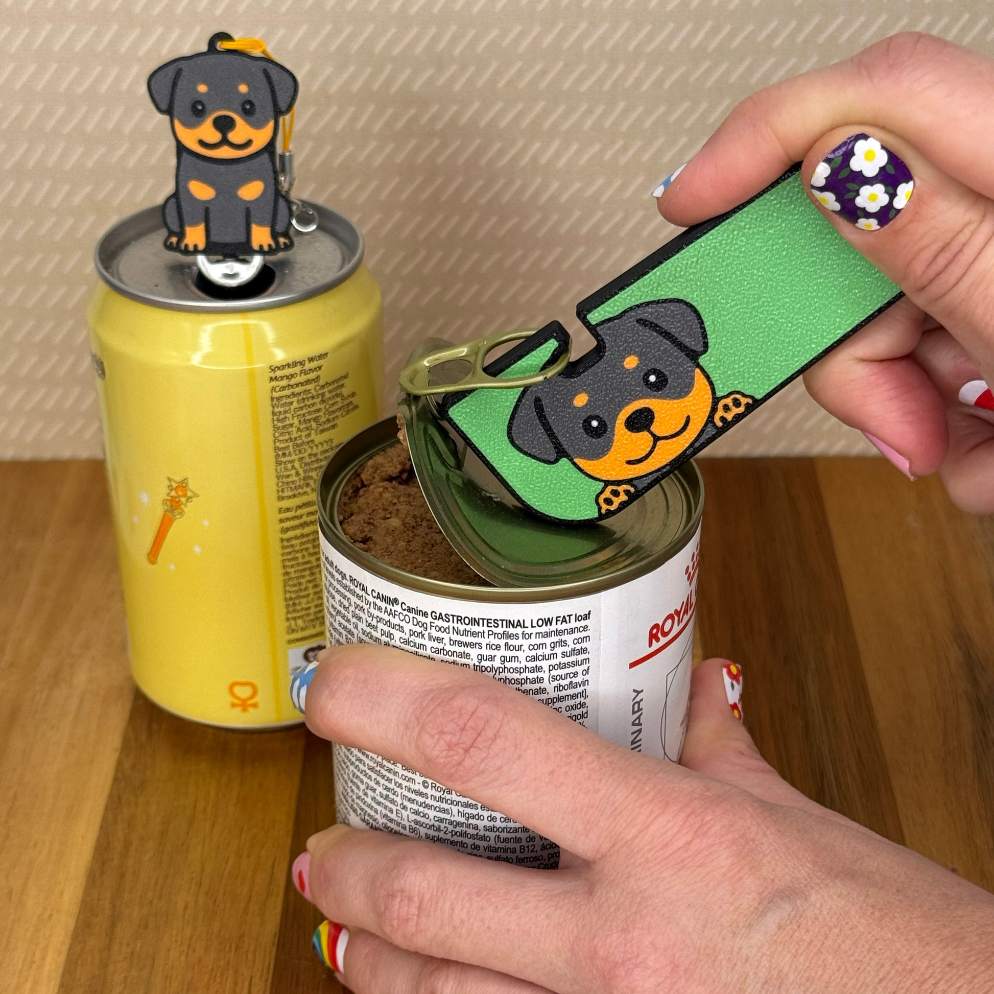 A person opens a can of dog food with a Rottweiler Dog-design Tab Buddy XL. In the background, a matching soda-sized Tab Buddy rests atop the tab of a can of sparkling water.