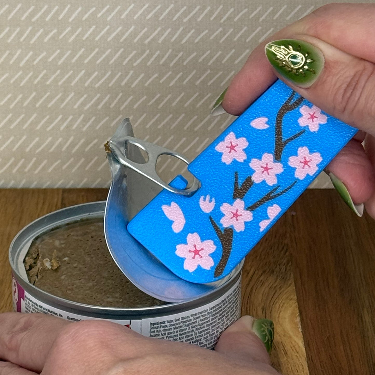 A Tab Buddy XL with a blue and pink cherry blossom design is used to peel back the tab lid on a can of cat food. 