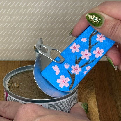 A Tab Buddy XL with a blue and pink cherry blossom design is used to peel back the tab lid on a can of cat food. 