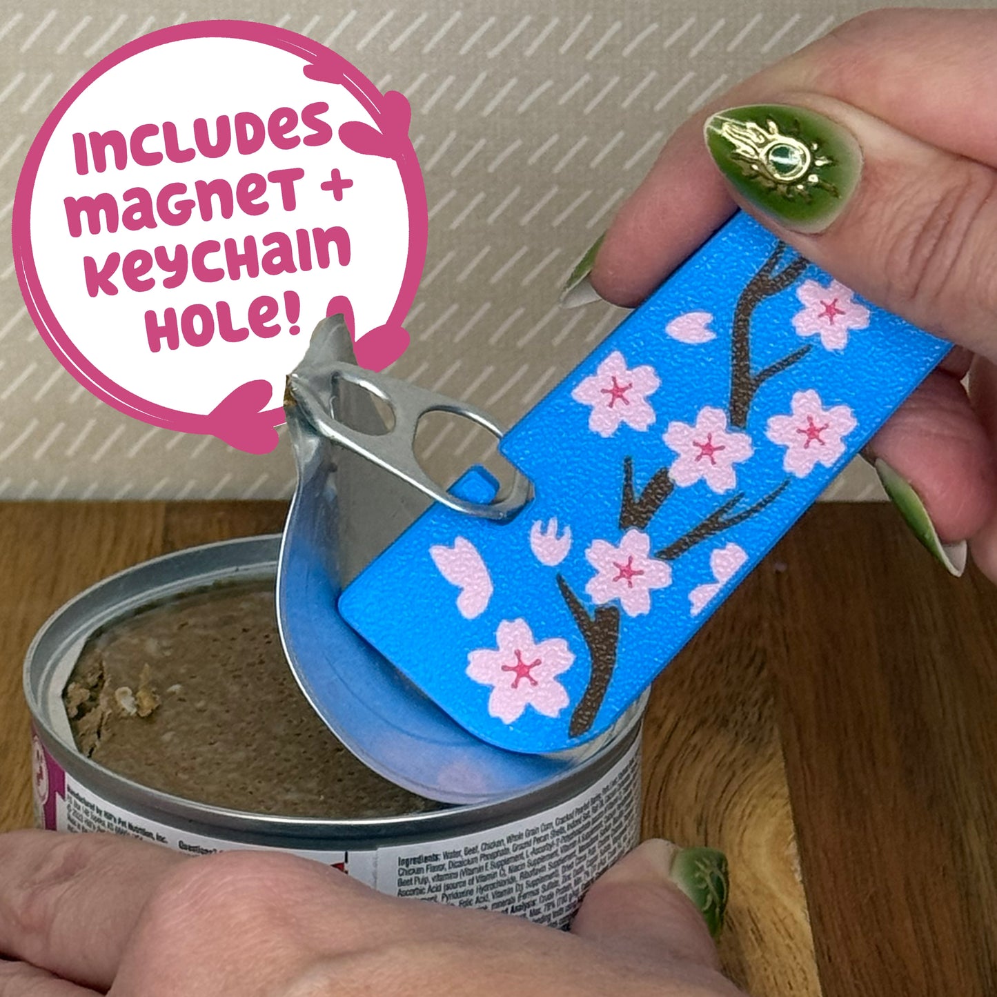 A Tab Buddy XL with a blue and pink cherry blossom design is used to peel back the tab lid on a can of cat food. Text reads "Includes magnet + keychain hole!"