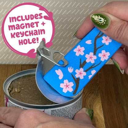 A Tab Buddy XL with a blue and pink cherry blossom design is used to peel back the tab lid on a can of cat food. Text reads "Includes magnet + keychain hole!"