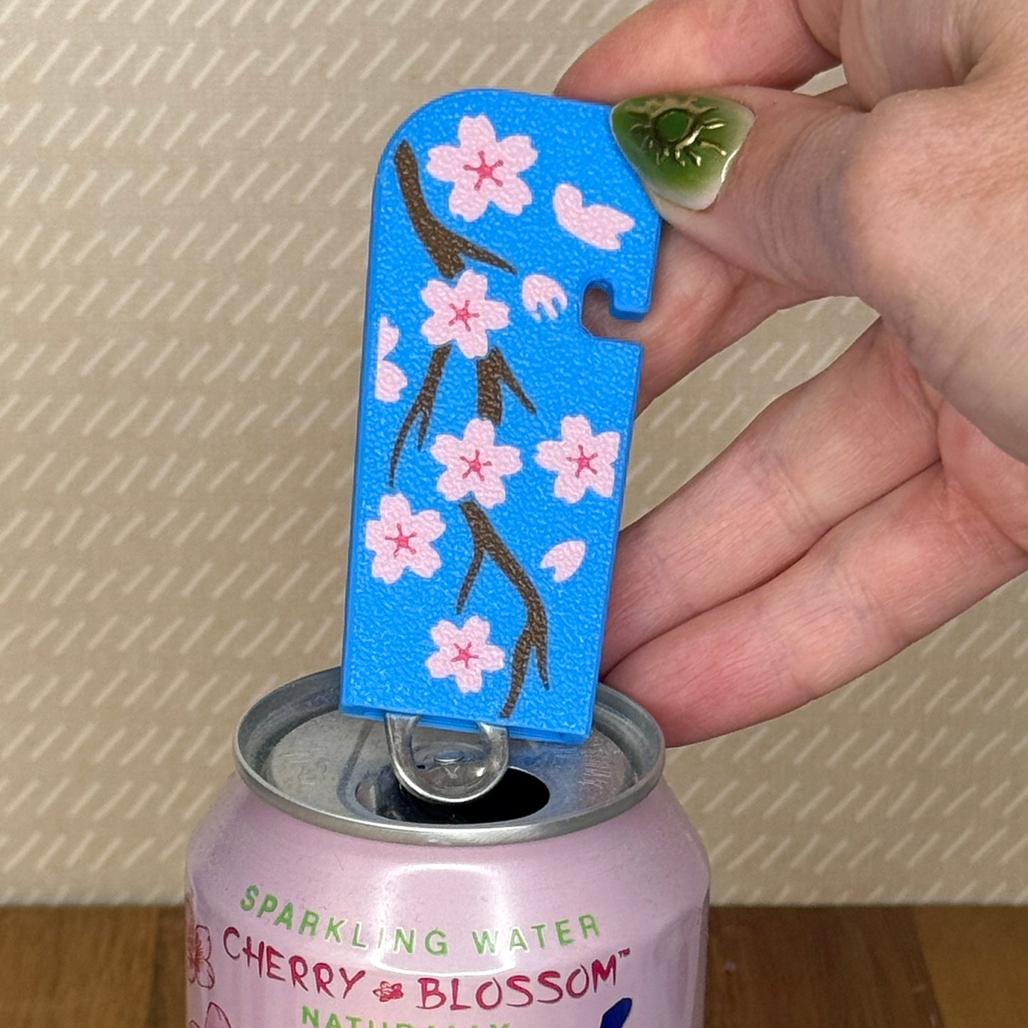 A cherry blossom design Tab Buddy XL is used to open a can of sparkling water  against a beige background.