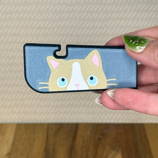 A sneaky cat Tab Buddy XL is held in a hand against a beige background. The cat is a blue-eyed ragdoll peeking up along the edge of the XL with a bluish-grey background.
