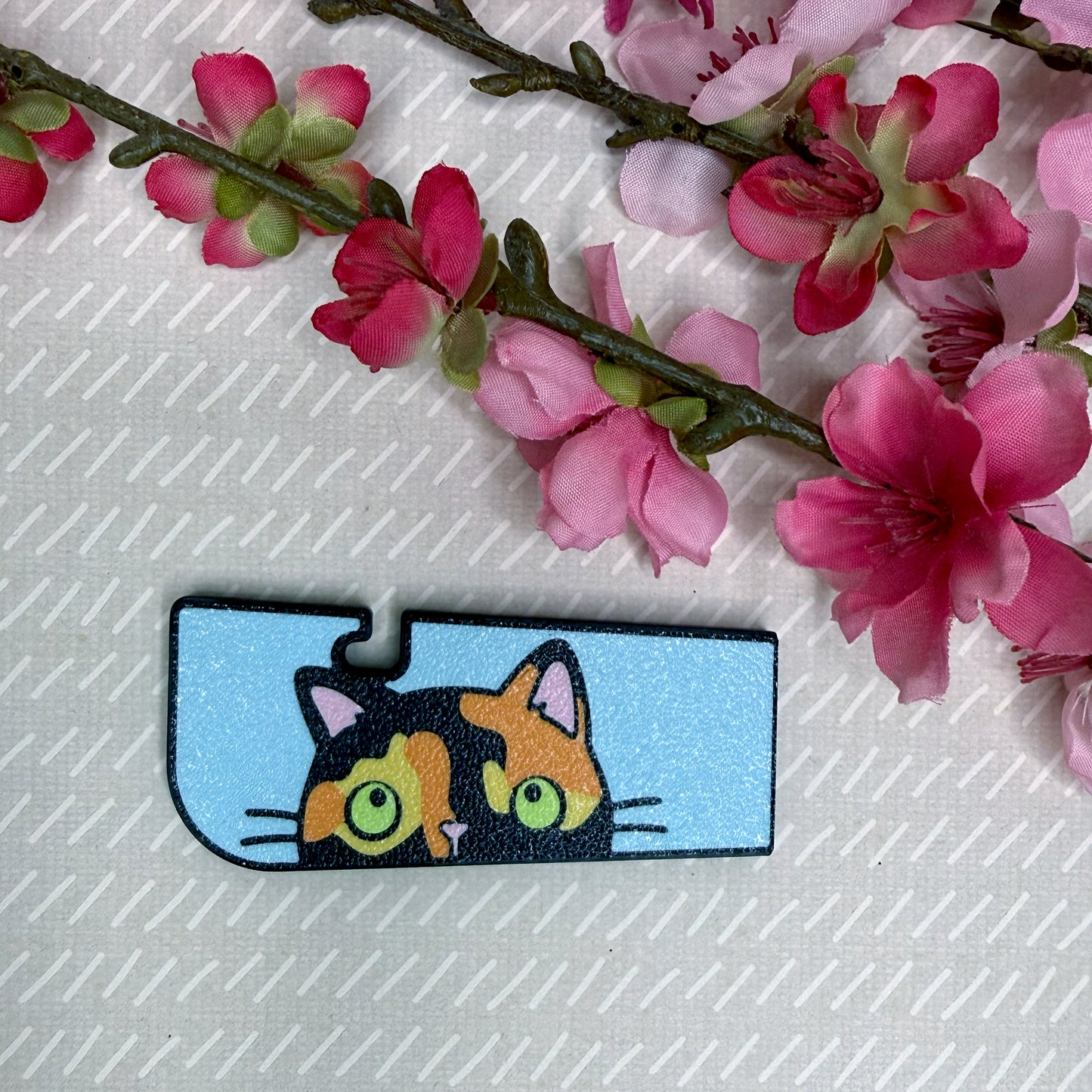 A Tab Buddy XL in Sneaky Tortoiseshell Cat color variation rests atop a beige surface with cherry blossom flowers in background.