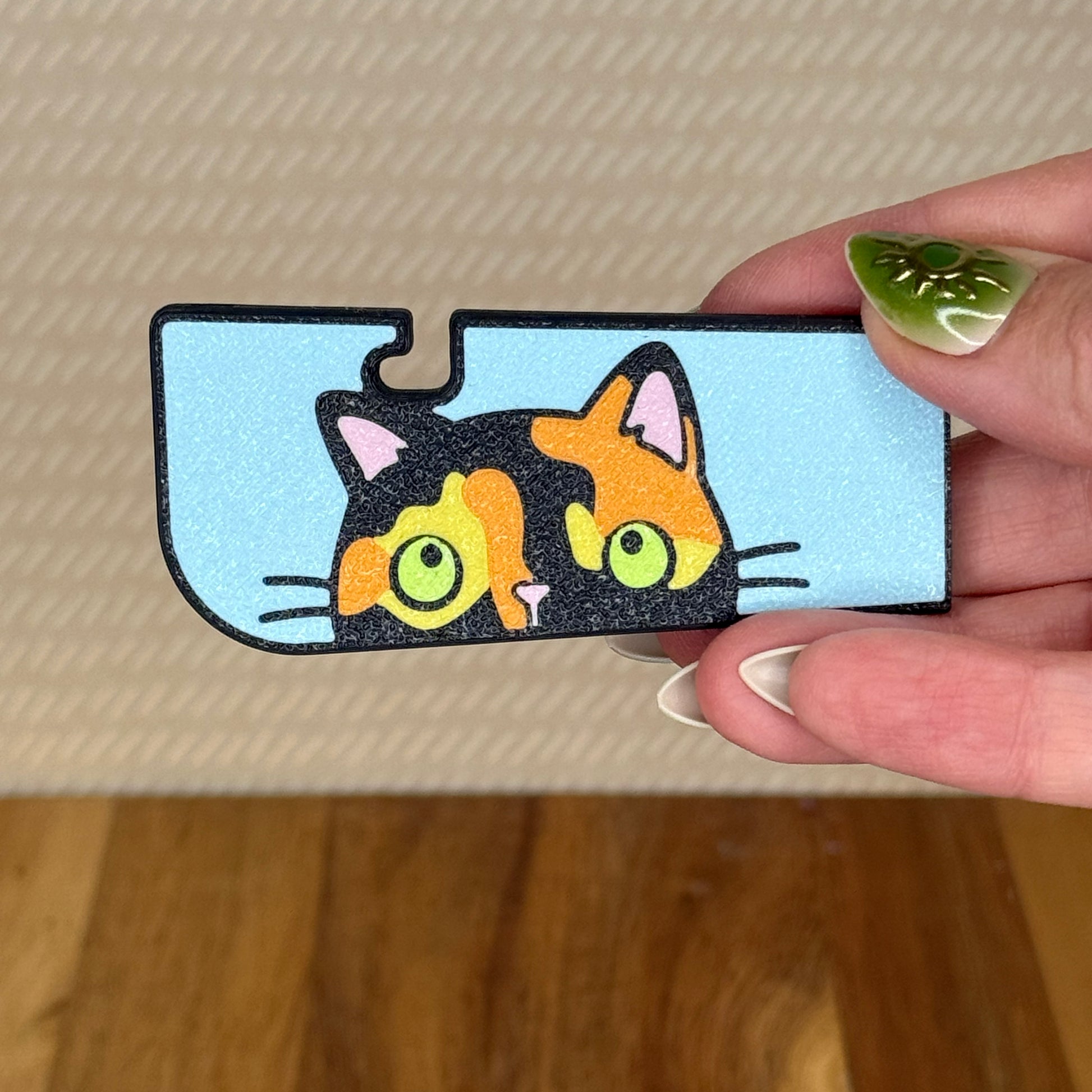 A sneaky cat Tab Buddy XL is held in a hand against a beige background. The cat is a green-eyed tortoiseshell peeking up along the edge of the XL with a blue background. 