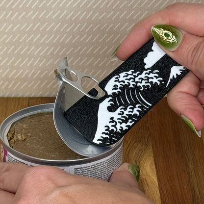A Tab Buddy XL with a black and white Great Wave design is used to peel back the tab lid on a can of cat food. 