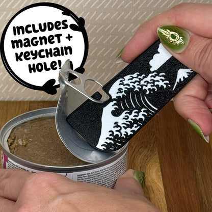 A Tab Buddy XL with a black and white Great Wave design is used to peel back the tab lid on a can of cat food. Text reads "Includes magnet + keychain hole!"