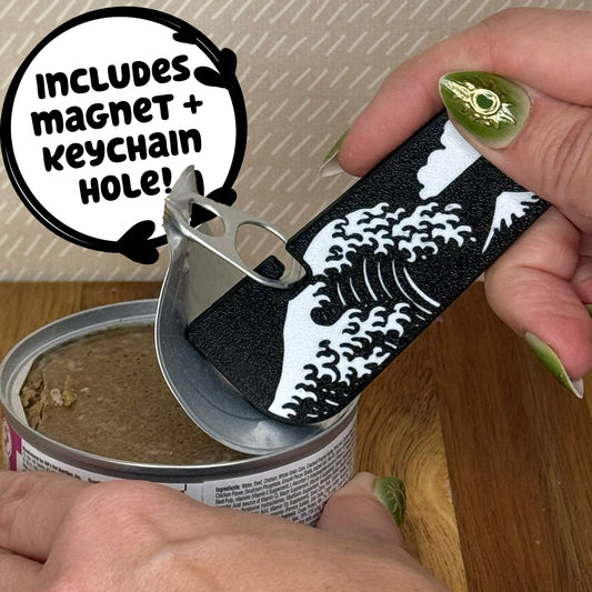 A Tab Buddy XL with a black and white Great Wave design is used to peel back the tab lid on a can of cat food. Text reads "Includes magnet + keychain hole!"