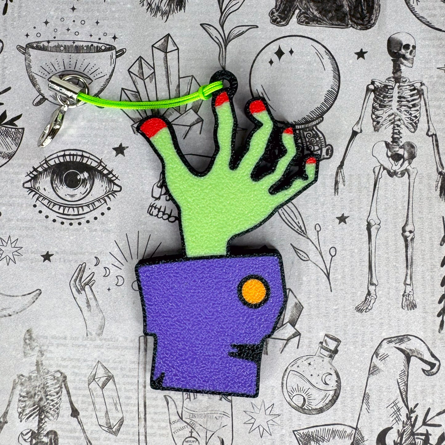 Green zombie hand Tab Buddy with red nails and keyring strap against a mystical background 